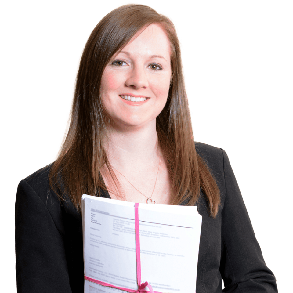 A woman with long browny red hair is smiling and holding a stack of papers tied with a pink ribbon.