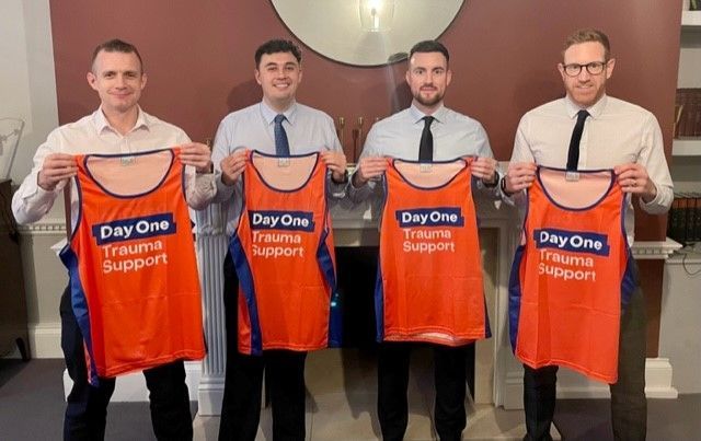 Four men in formal attire hold up orange and blue "Day One Trauma Support" jerseys while standing indoors.